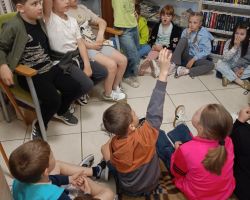 Dzieci siedzą na podłodze w bibliotece i zgłaszają się do odpowiedzi.