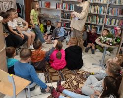 Uczniowe siedzą na podłodze w bibliotece i słuchają bibliotekarki.