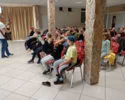 Uczniowie siedzą na krzesłach i oglądają mężczyznę, który stoi i gra na gitarze.