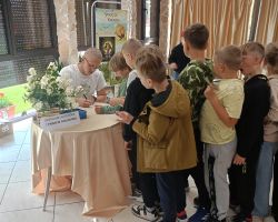 Dzieci stoją w kolejce po autograf od autora książek, który siedzi przy stoliku na krzesełku.