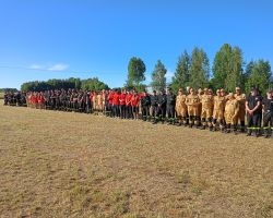 Drużyny strażackie, kobiety i mężczyźni, stoją ustawieni w równym rzędzie.