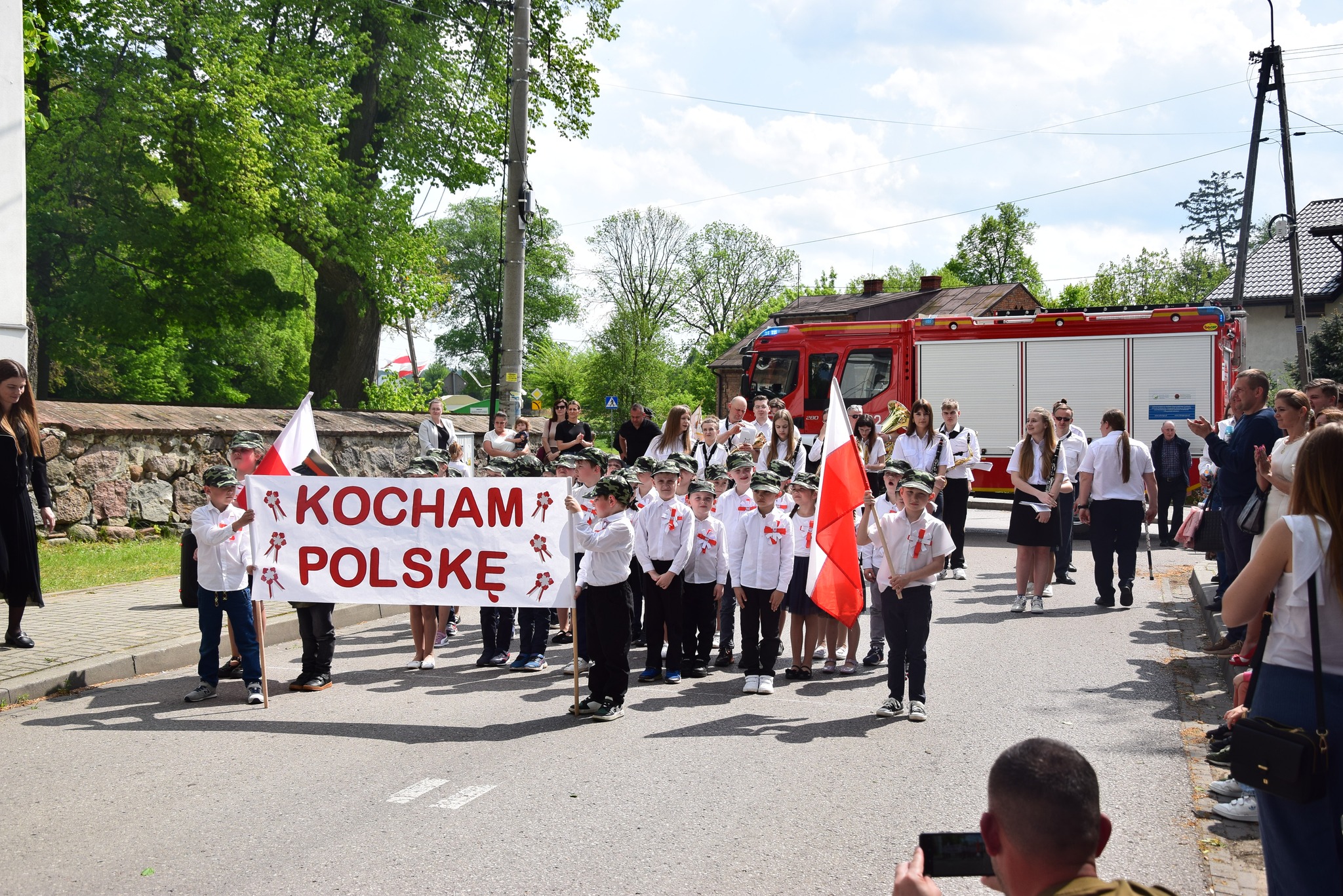 Grupa dzieci ubrana na galowo trzyma w rekach flagi i transparent Kocham Polskę.